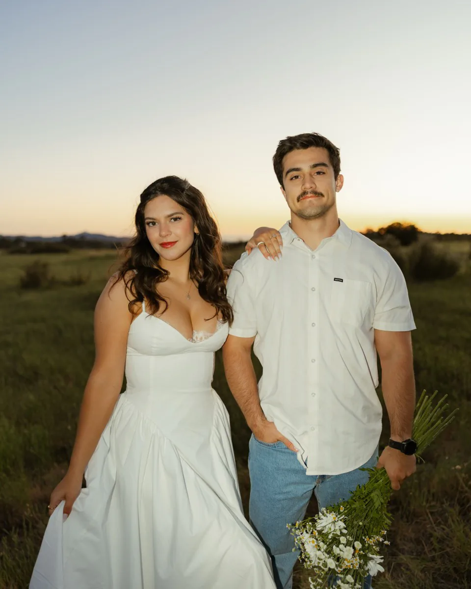 A couple photographed by Brooke Bell Photography: at sunset in an open field, she in a white dress holding her skirt, he holding a bouquet of wildflowers, both faces to the camera