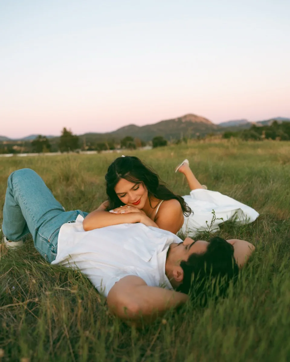 The same couple lying in tall grass at dusk, mountains behind them