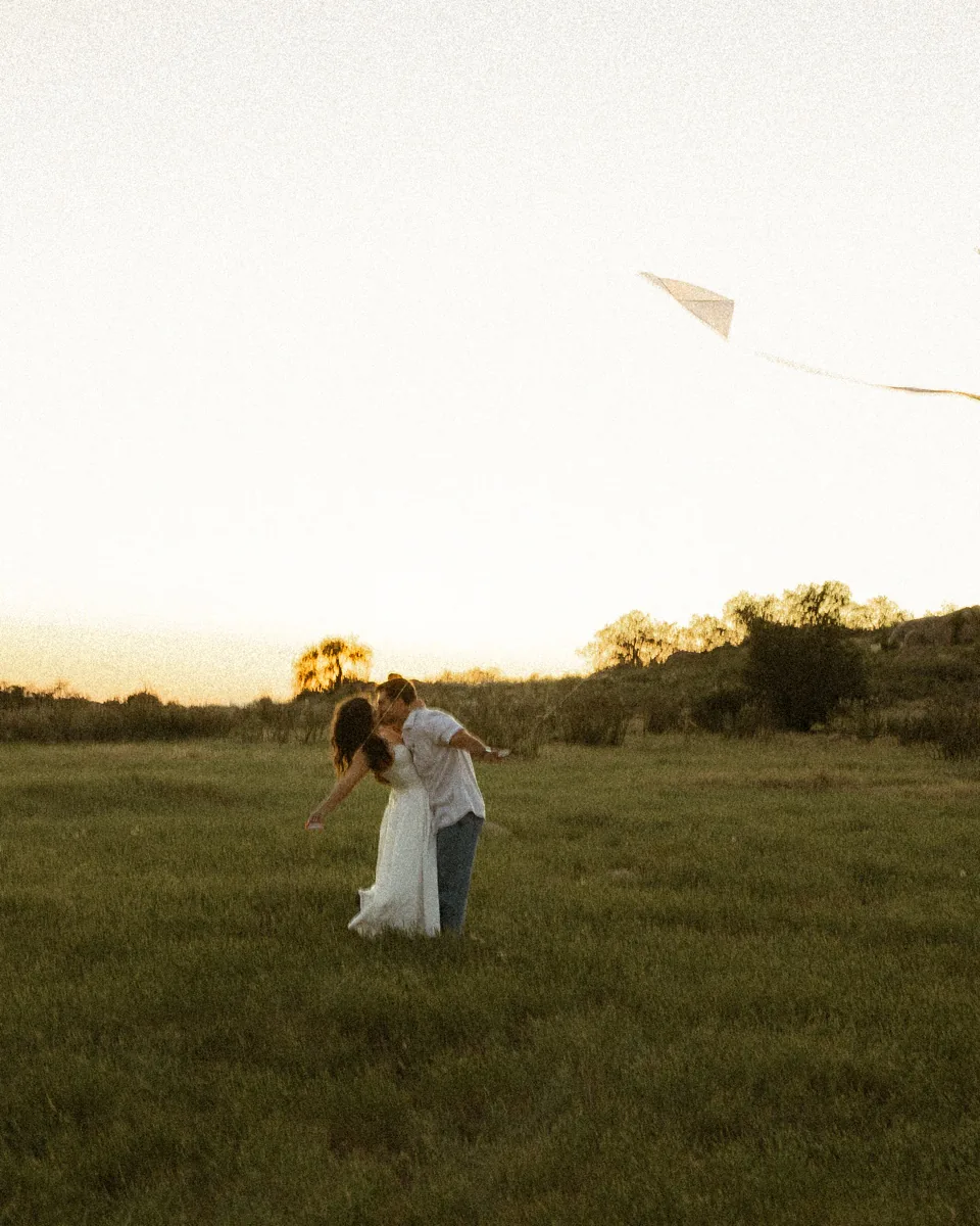 The couple kissing in a wide field while two kites fly overhead